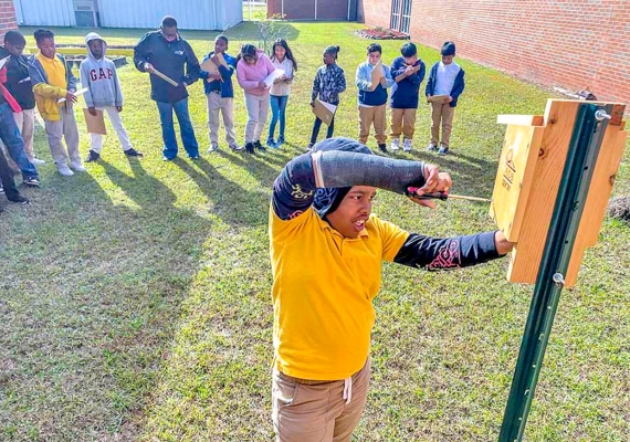 Student works on birdhouse while other students watch in the background.