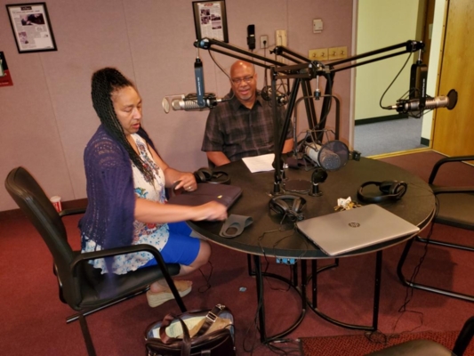 Two people sit in a radio studio in front of microphones.